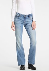 Personne portant une chemise blanche ajustée à manches longues, un jean bootcut bleu clair et des bottes noires, debout devant un fond uni clair.