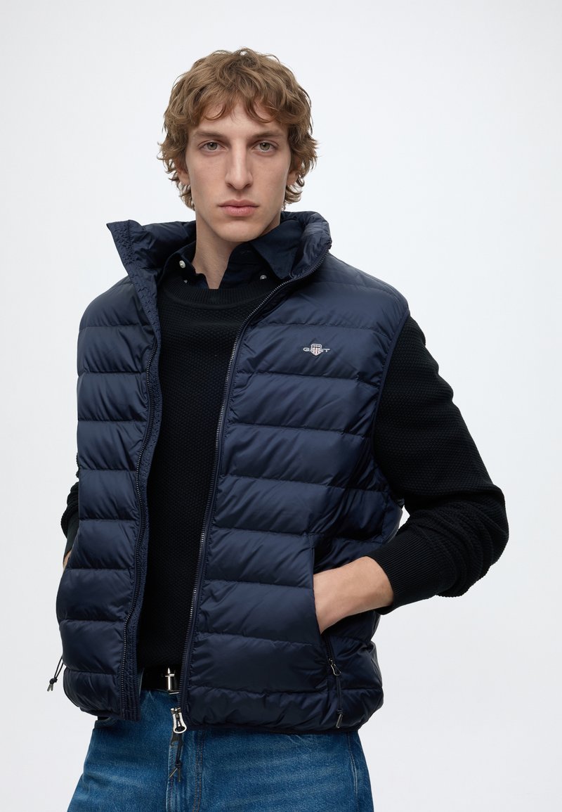 Ung mand med krøllet hår iført en marineblå quiltet vest over en sort sweater og blå jeans, stående med hænderne i vestens lommer.