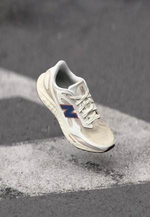 Scarpa da corsa New Balance beige chiaro e bianca con logo rosso-blu, sospesa sopra l'asfalto con segni di frecce bianche.