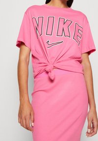 Rosa Nike-t-shirt med korta ärmar, med stor svart text. T-shirten är knuten i midjan och matchas med en rosa ribbad kjol.