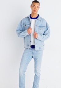 Joven con chaqueta de mezclilla azul clara y jeans, ajustando el cuello de la chaqueta, de pie contra un fondo blanco liso.