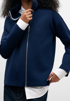 Person iført en mørkeblå jakke med lynlås over en hvid skjorte, med den ene hånd, der justerer kraven, og den anden i lommen, mod en ensfarvet baggrund.