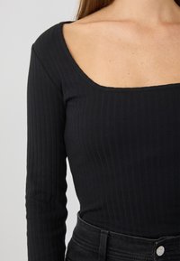 Schwarzes, geripptes Langarmoberteil mit quadratischem Ausschnitt. Hat ein figurbetontes Design und eine glatte Textur. Kombiniert mit hochgeschnittenen schwarzen Jeans.