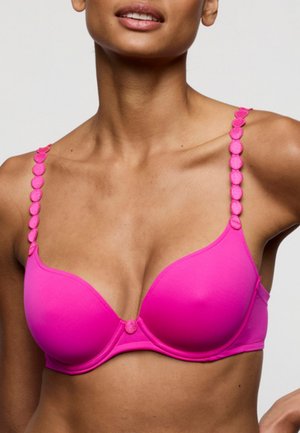 Vrouw draagt een felroze beha met decoratieve ronde details op de bandjes en in het midden vooraan, getoond van schouders tot halverwege de torso.