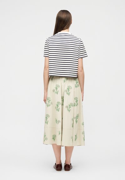 Γυναίκα με την πλάτη, φοράει κοντομάνικη μπλούζα με λευκές και ναυτικό-μπλε ρίγες, κρεμ φούστα με πράσινα λουλουδένια σχέδια και καφέ παπούτσια.