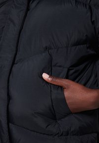 Puffjacka i svart nylon med quiltat mönster. Har en sidoficka, med en delvis synlig hand som vilar på tyget.
