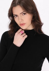 Rózsaszín gumi gyűrű, texturált mintával az mutatóujjon, ellentétben egy fekete bordázott garbóval, közepes hosszúságú, hullámos hajjal.