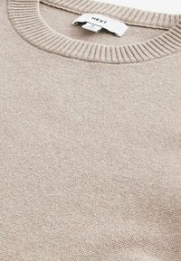 Beżowy sweter z okrągłym dekoltem, ściągaczami na rękawach i na dole, teksturowanym wzorem dzianiny oraz małą metką przy szyi wskazującą markę.