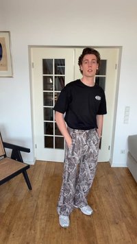 Fiatal férfi áll bent, kezét egy mintás, bő szárú nadrág zsebében tartja, fekete pólót és fehér tornacipőt visel fa padlón.