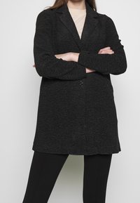 Blazer noir texturé avec une fermeture à un bouton, des revers crantés et des manches longues, porté sur un haut clair et un pantalon noir.