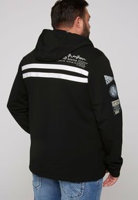Chaqueta negra con capucha que presenta rayas horizontales blancas, logos bordados y parches en las mangas. Textura suave con un ajuste relajado.