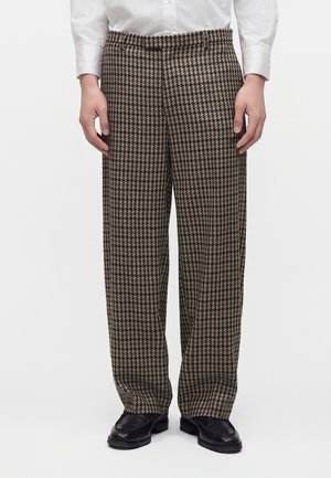 Personne portant un pantalon large à motif pied-de-poule marron et noir, une chemise blanche et des chaussures noires, debout devant un fond blanc uni.