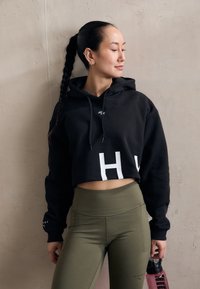 Felpa nera corta con grande lettera bianca "H" e cordoncino, abbinata a leggings verdi oliva a vita alta, mentre tiene in mano una bottiglia d'acqua rosa.