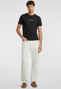 Juodas medvilninis marškinėliai su balta „GUESS JEANS“ logotipu, derinami su grietinėlės spalvos džinsais ir rudos zomšinės avalynės. Įprastas kirpimas, trumpomis rankovėmis.