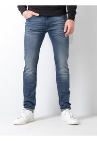 Jeans de mezclilla azul con un corte ajustado, que presentan áreas desgastadas, diseño de cinco bolsillos y bordes desgastados. Combinados con zapatillas blancas.