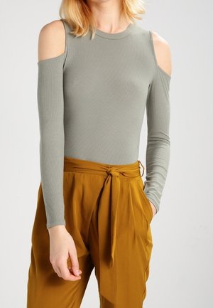 Vrouw met een geribbelde, lange mouwen, grijze cold-shoulder top, gecombineerd met hooggetailleerde mosterdgele broek met een strik in de taille.
