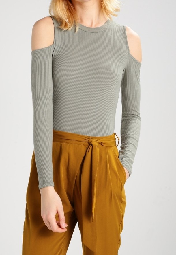 Femme portant un haut gris côtelé à manches longues et épaules dénudées, associé à un pantalon taille haute moutarde noué à la taille.