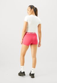 Fehér, testhez simuló póló és rózsaszín rövidnadrág, az övén a "NIKE" logóval, sima textúrával, fekete sportcipővel és fehér zoknival párosítva.