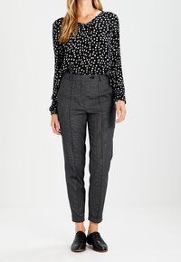 Blouse à pois noirs et blancs avec manches longues, associée à un pantalon à carreaux gris à coupe fuselée et plis avant. Chaussures noires.