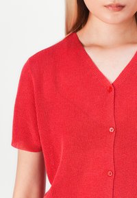 Rød kortermet cardigan med v-hals, vevd tekstur og to frontknapper. Minimalistisk design uten ytterligere mønstre eller aksenter.