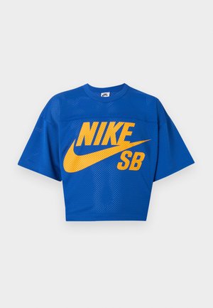 Jeune homme portant un t-shirt en mesh bleu Nike SB, un jean en denim bleu clair et une casquette Nike bleu clair, debout devant un fond uni.