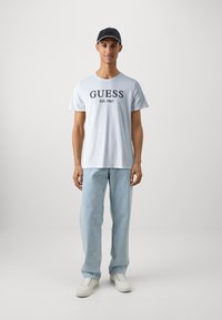 Bílá bavlněná tričko s potiskem "GUESS EST. 1981" v černé barvě, v kombinaci s světle modrými džíny a bílými teniskami, doplněné tmavou čepicí.