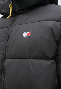 Jachetă puffer neagră cu fermoar frontal, având un șnur galben, textură matelassé și un logo multicolor Tommy Jeans pe piept.