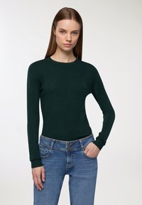 Ciemnozielony dopasowany sweter z długimi rękawami i okrągłym dekoltem, w połączeniu z niebieskimi dżinsami z guzikiem i kieszeniami.