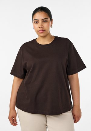 Ung kvinde med mørkt hår sat tilbage, iført en løs mørkebrun kortærmet skjorte og beige bukser mod en ensfarvet baggrund.