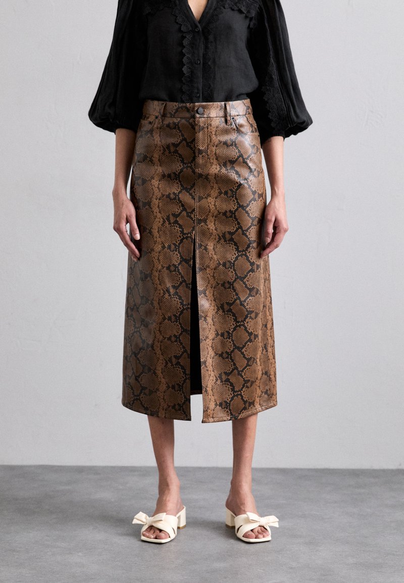 Bruine leren midi-rok met slangenpatroon en een split aan de voorkant, gecombineerd met crème blokhak sandalen en een zwarte blouse met knopen.