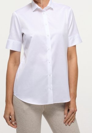 Donna che indossa una camicia bianca a maniche corte e pantaloni beige chiaro con trama, in piedi con una mano rilassata al fianco.
