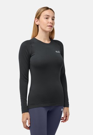 Frau trägt ein enganliegendes schwarzes langärmliges Sportoberteil und blaue Leggings und steht vor einem schlichten hellen Hintergrund.