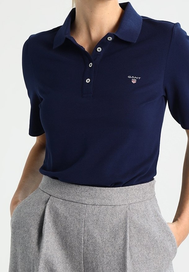 Femme portant un polo bleu marine à manches courtes avec des boutons et un pantalon gris plissé taille haute, mains partiellement dans les poches.