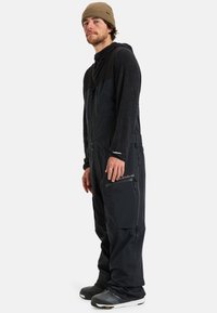 Czarne ocieplane ogrodniczki z zamkiem błyskawicznym z przodu, kieszeniami bocznymi i fakturowaną górną częścią. Noszone z dopasowanym ciemnym swetrem i czapką beanie.