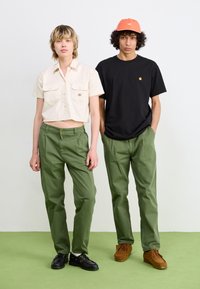 Two models wear casual outfits: a cropped cream button-up shirt, olive green pants, and black shoes; alongside a black t-shirt, olive pants, and brown shoes.