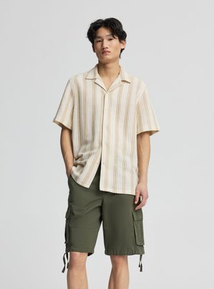 Giovane uomo in piedi con le mani in tasca, indossa una camicia beige a righe maniche corte e pantaloncini cargo verde oliva su sfondo semplice.