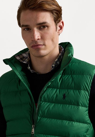 Green padded vest made of shiny material, featuring a high collar, zip closure, and a small logo. Worn over a dark sweater and plaid shirt.