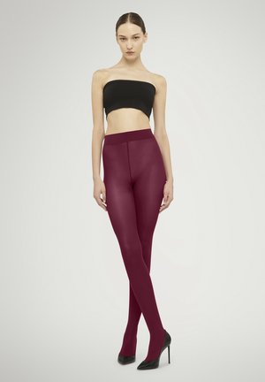 Kvinde stående med krydsede ben iført en sort tube-top, bordeauxugennemsigtige strømpebukser og sorte højhælede sko på en ensfarvet baggrund.