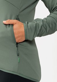 Haut vert à manches longues avec une finition texturée, doté d'une poche zippée et d'un accent contrastant. Le tissu est lisse et ajusté.