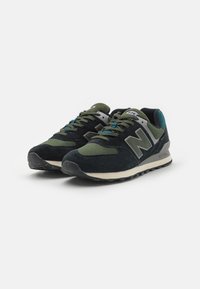 Černé a olivově zelené tenisky New Balance 574 s svrchním materiálem z semiše a síťoviny, zahrnující šedé logo "N" a design na šněrování, bílou mezipodešev a černou podrážku.