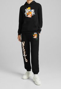 Sudadera negra con estampado gráfico de un personaje en el frente; pantalones sudadera a juego con gráfico del personaje y texto "NARUTO" en naranja. Zapatillas blancas.