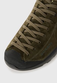 Chaussure en suede vert foncé avec un embout en caoutchouc, des lacets plats et des coutures renforcées. Marquage visible sur le côté.