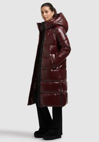 Hosszú, bordó, fényes hatású tollkabát, túlméretezett kapucnival, patentos gombokkal, oldalzsebekkel és steppelt mintázattal. Fekete részletek a cipzárokon.