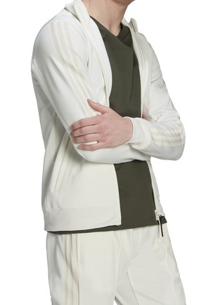 Veste blanche zippée avec capuche, ornée de rayures crème clair sur les épaules. Assortie à un t-shirt col rond vert olive. Texture lisse.