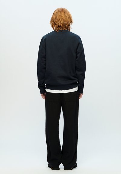 Sweatshirt bleu marine foncé avec un col rond, des poignets et un ourlet côtelés, superposé sur une chemise blanche. Assorti avec un pantalon large noir.