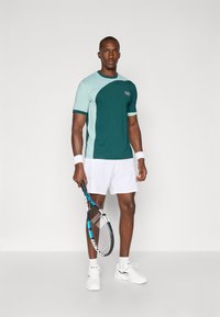 Tennisantrekk med en blågrønn og myntefarget t-skjorte med buet design, kombinert med hvite shorts og hvite joggesko, holdende en racket.