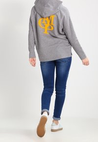 Grijze hoodie met een geel geborduurd logo, relaxed fit, met een voorzak in kangoeroestijl. Gecombineerd met donkerblauwe jeans en witte sneakers.