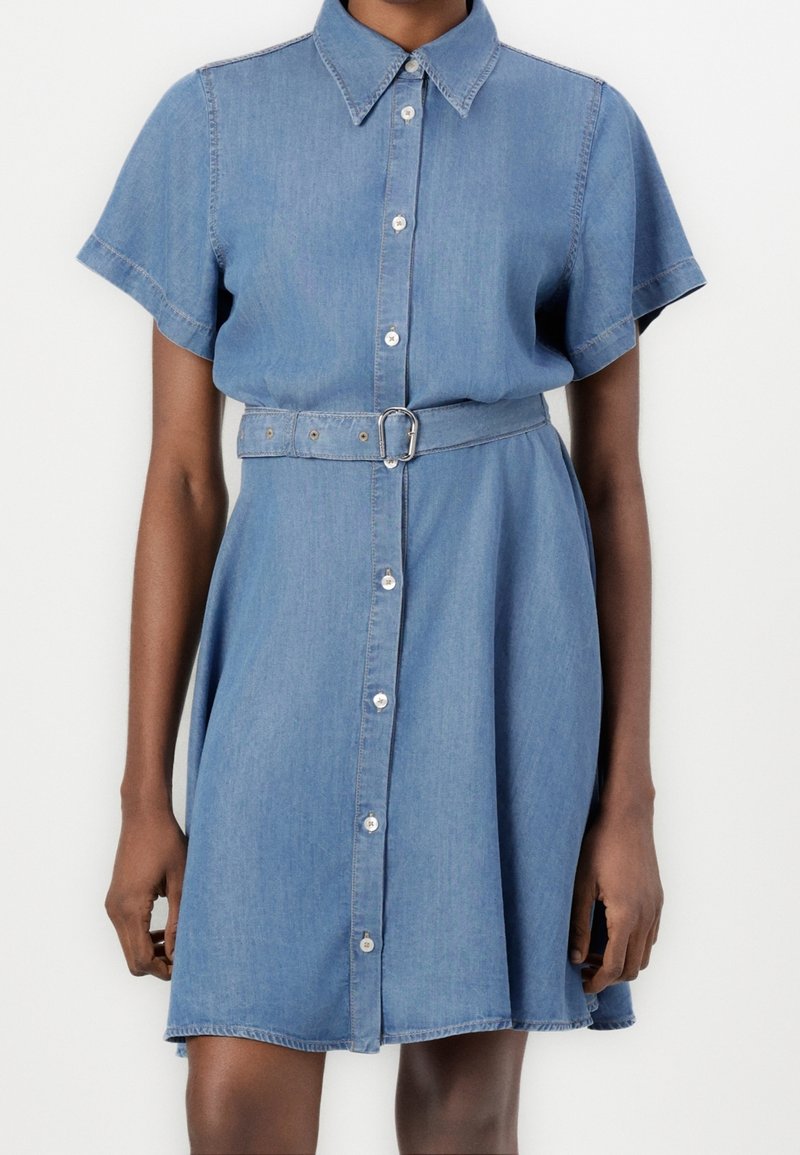 Robe en jean bleue à manches courtes avec des boutons sur le devant et une ceinture assortie à la taille, portée par une personne debout sur un fond blanc.
