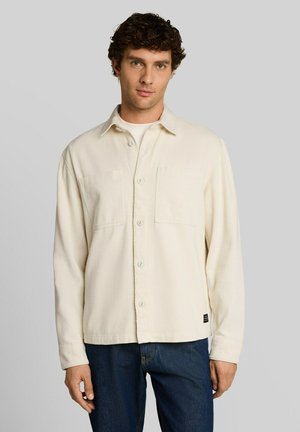 Junger Mann mit lockigem dunklem Haar, der ein cremefarbenes Hemd über einem weißen Top und dunkelblauen Jeans trägt, steht vor einem einfarbigen grauen Hintergrund.