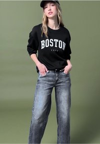 Sudadera negra con "BOSTON 1900" en blanco, combinada con jeans de mezclilla gris claro. El modelo lleva una gorra. El fondo es verde.